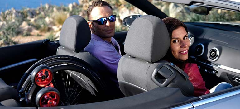 A man and woman in a convertible with a wheelchair in the backseat